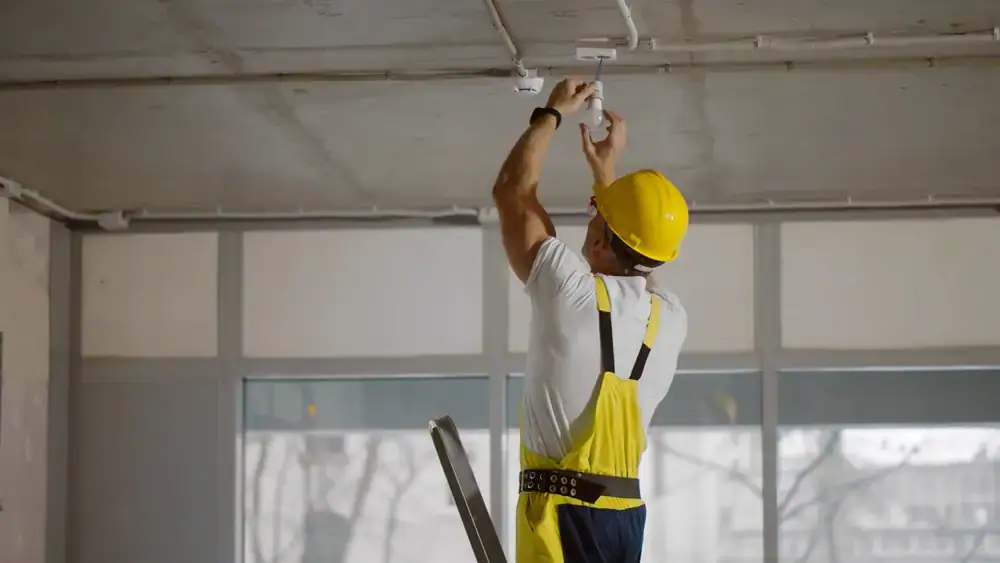 An electrician installing a ceiling light fixture in a home in Chicago, IL. The technician is using a ladder and wearing safety gear while connecting wiring to the ceiling.