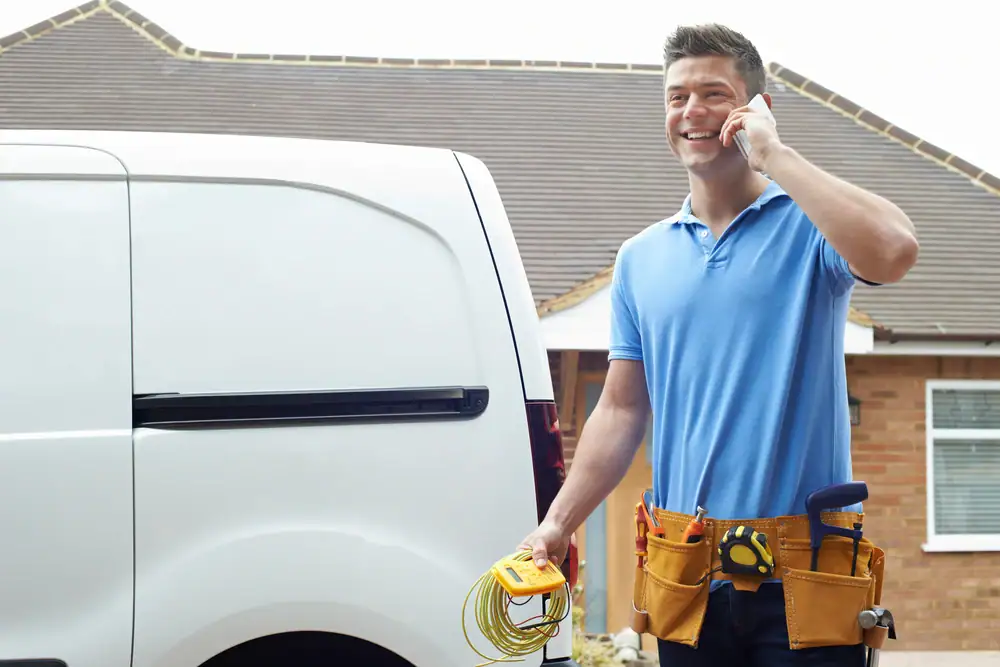 A smiling electrician Cook and Will County wearing a tool belt holds wires and talks on a smartphone while standing next to a white van outside a house in IL.