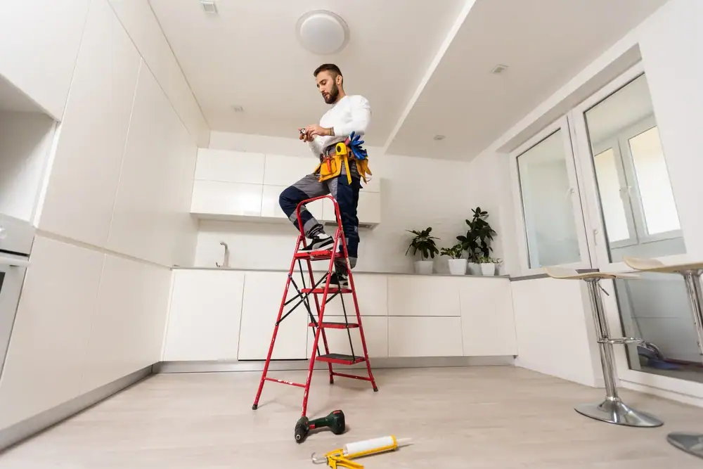 A man, likely an electrician in Cook and Will County, stands on a red ladder in a white kitchen, wearing a tool belt and working with electrical wires. Tools are scattered on the floor beneath him.