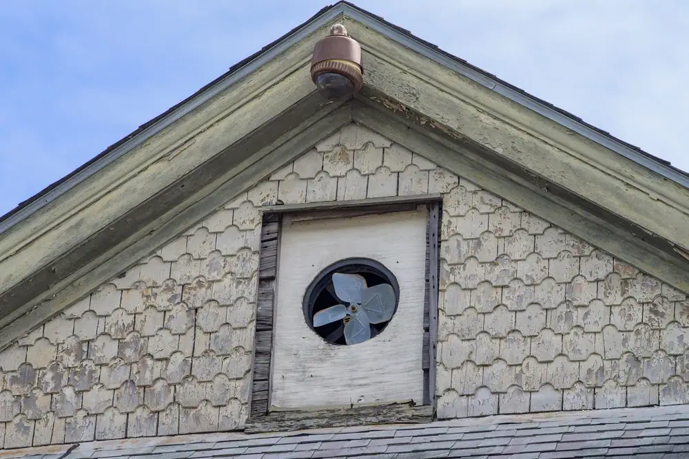 Weathered building gable with peeling paint, a circular vent featuring a fan in the center, and an old security camera at the roof peak against a blue sky—a common sight for an electrician Cook and Will County, IL, restoring electrical services.