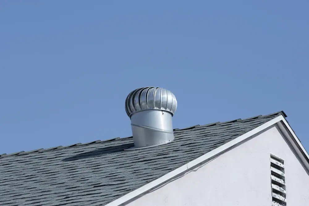 A close-up of a gray attic turbine vent installed on the sloped shingle roof of a building in IL, set against a clear blue sky—perfect for homes serviced by electrician Cook and Will County.