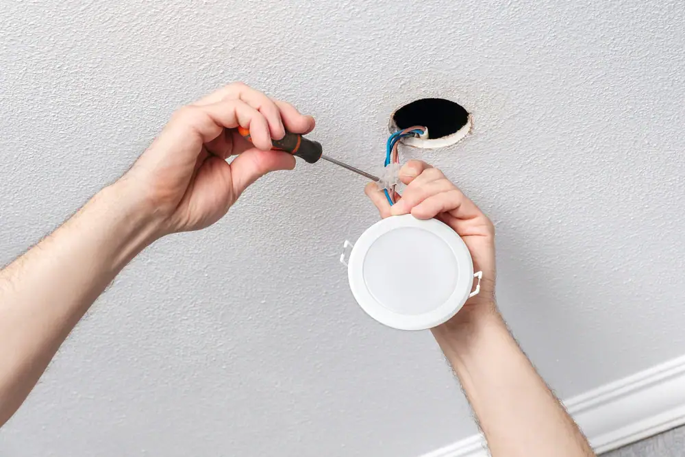 An electrician in Cook and Will County uses a screwdriver to install or wire a round recessed ceiling light, with blue and brown wires visible from a hole in the white textured ceiling—expert electrical services for your IL home.
