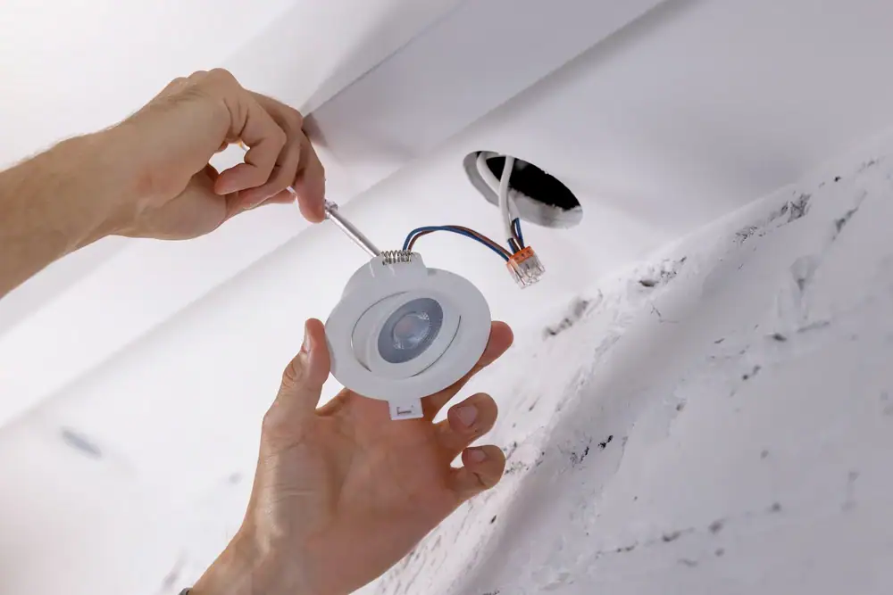 Close-up of hands installing a recessed ceiling light fixture, with exposed wires visible and a white, unfinished ceiling in the background—expertly handled by an electrician Cook and Will County, IL.