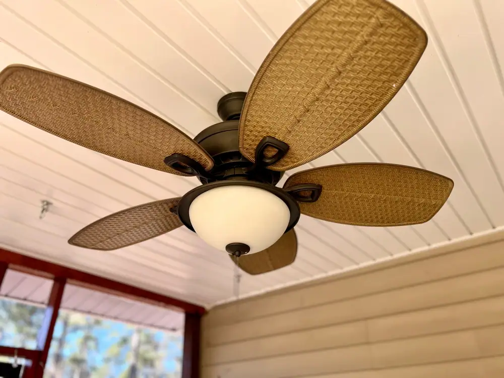 A ceiling fan with five brown, woven-texture blades and a central white light fixture is mounted on a white paneled ceiling in a room with beige siding—expertly installed by an electrician in Cook and Will County, IL.
