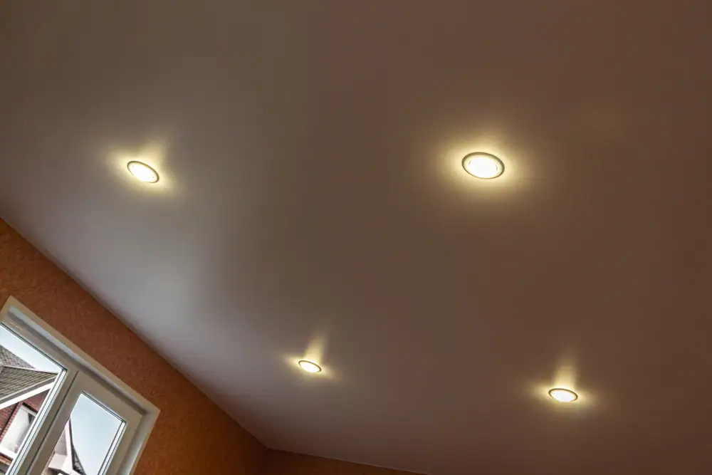 A ceiling with four recessed lights installed by an electrician Cook and Will County, casting soft glows. Part of a window and the upper corner of the room are visible. The walls are painted a warm color.