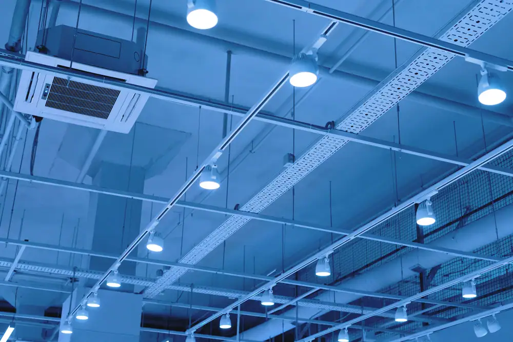 A modern ceiling with exposed pipes, vents, wiring trays, and multiple round LED lights illuminated by blue-tinted light—perfectly showcasing quality Electrical Services in Cook and Will County, IL.