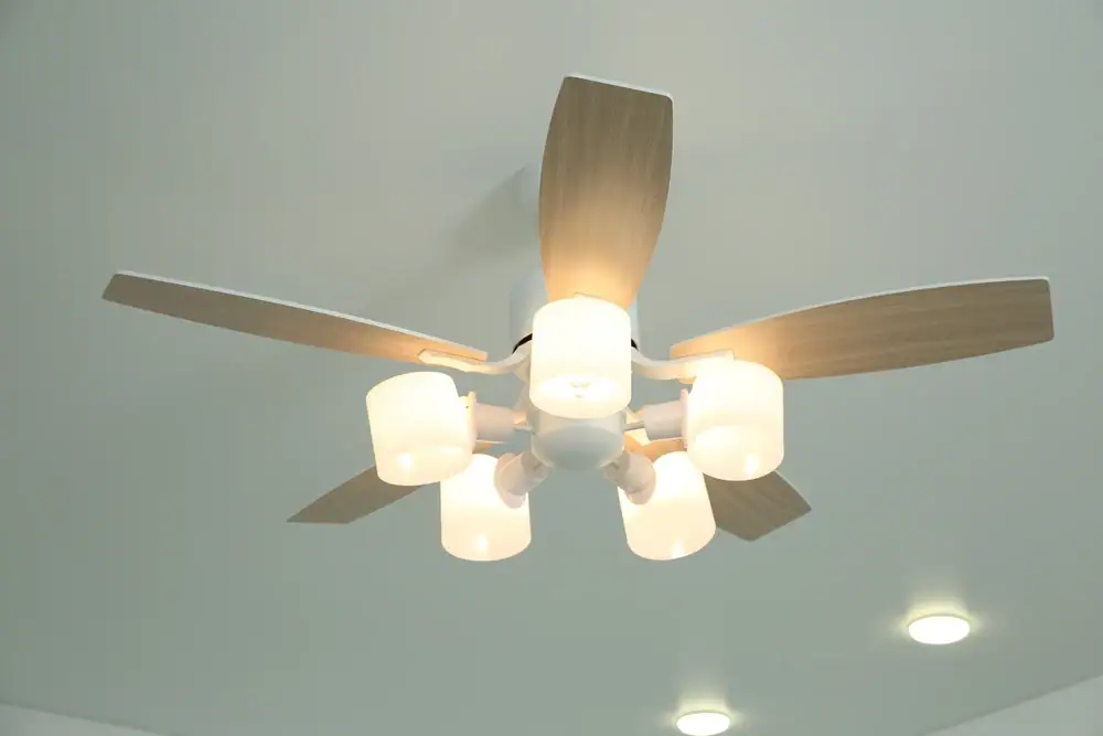 A ceiling fan with five light wooden blades and five attached white lampshades is mounted on a white ceiling. Two recessed ceiling lights are visible nearby, perfect for showcasing expert Electrical Services in Cook and Will County, IL.