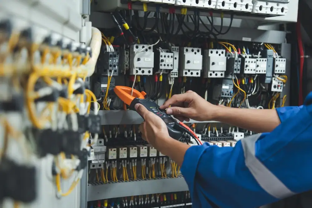 An electrician in Cook and Will County, IL, wearing a blue uniform, uses a clamp meter to check the wiring and components inside an open electrical control panel filled with wires and switches.