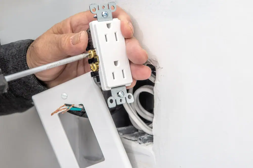 An electrician in Cook and Will County installs a white electrical outlet, using a screwdriver to secure the wires. The outlet cover plate and exposed wires are visible next to the open outlet box, showcasing professional Electrical Services in IL.