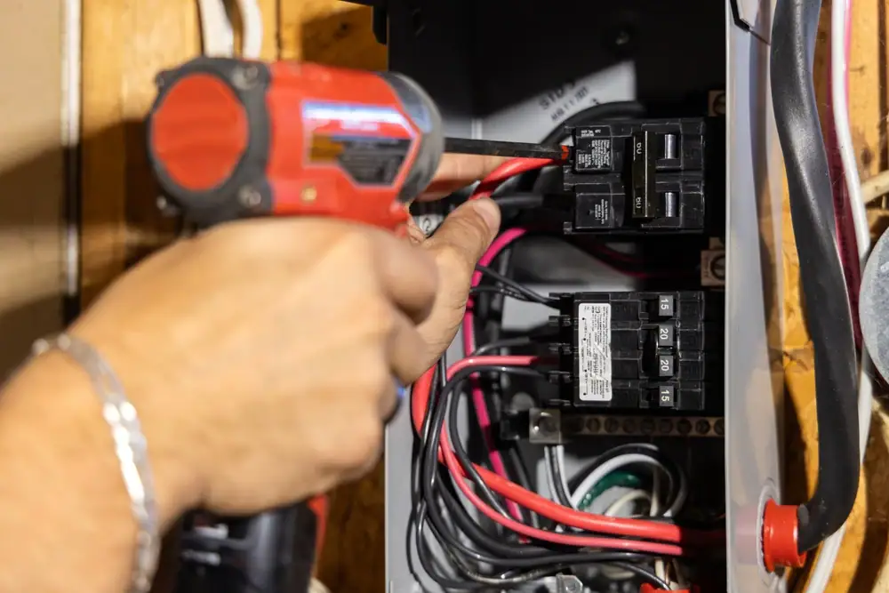 An electrician Cook and Will County, IL uses a cordless power drill near circuit breakers inside an open electrical panel, holding a red wire among various colored wires.