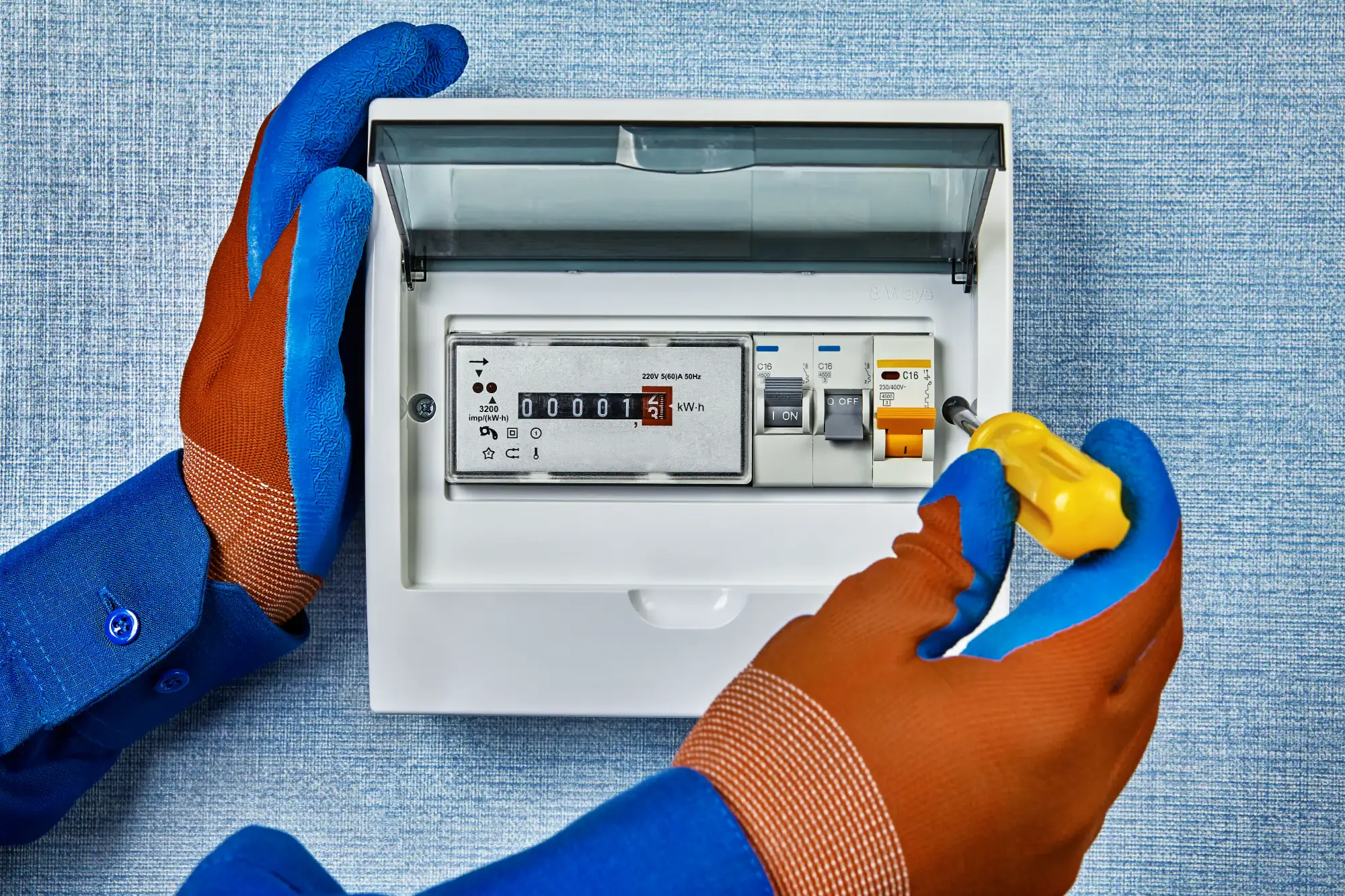 An electrician Cook and Will County, IL, wearing blue and orange protective gloves uses a yellow screwdriver to work on an electric meter box mounted on a wall.
