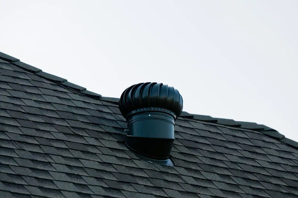 A close-up view of a black roof turbine vent installed on a shingled rooftop against a pale sky background, highlighting quality Electrical Services in Cook and Will County, IL.