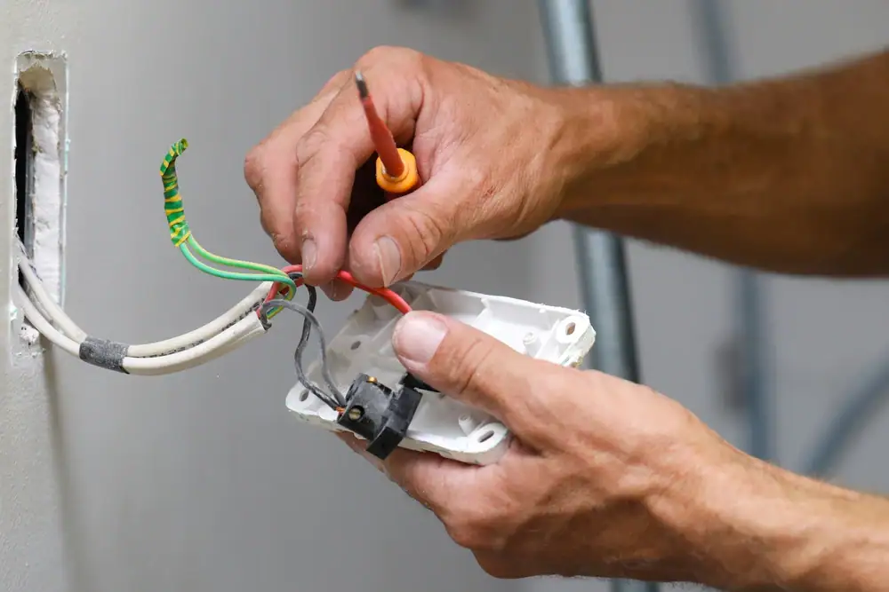 An electrician Cook and Will County, IL uses a screwdriver to work on an electrical outlet, connecting colored wires. The outlet cover is removed, exposing the internal wiring against a white wall.