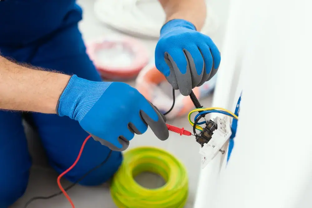 An electrician in Cook and Will County, IL, wearing blue gloves uses a multimeter to test electrical wires connected to a wall socket, with a coil of yellow wire nearby.