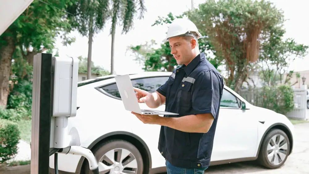 A worker wearing a white hard hat uses a laptop while inspecting an electric vehicle charging station for Electrical Services Cook and Will County, with a white car parked nearby and trees in the background.