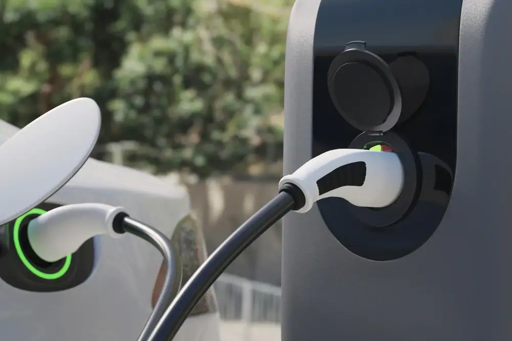Close-up of an electric car being charged, with a white charging cable plugged in. Electrical Services Cook and Will County ensure safe installs. A white car with a green ring on the port is parked nearby; blurred trees fill the background.
