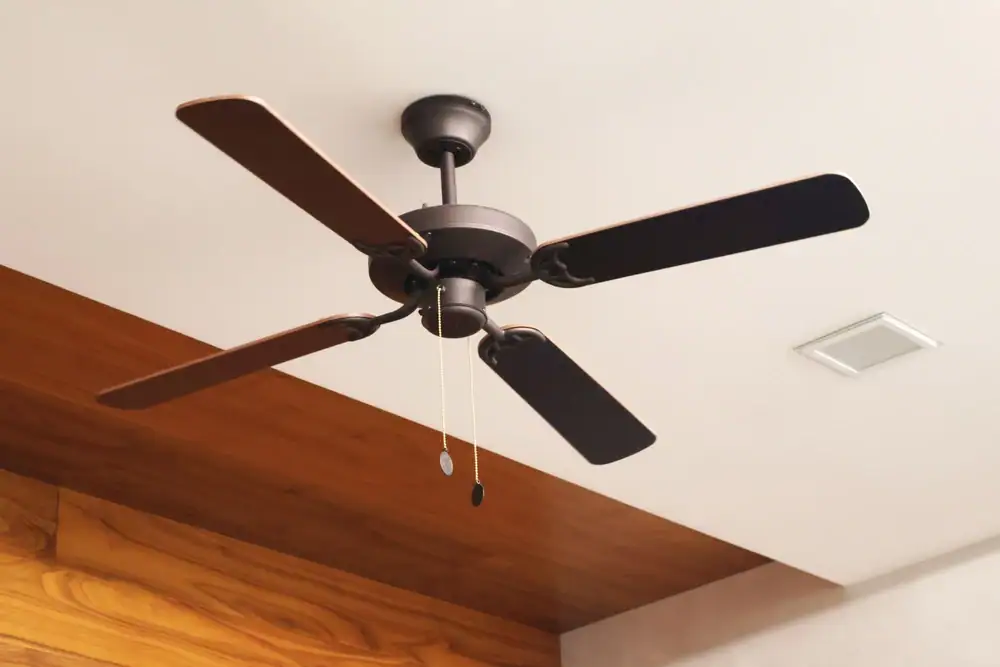 A four-blade ceiling fan with brown blades and pull chains is mounted on a white ceiling with a wooden accent and a square air vent nearby, expertly installed by an electrician Cook and Will County, IL.