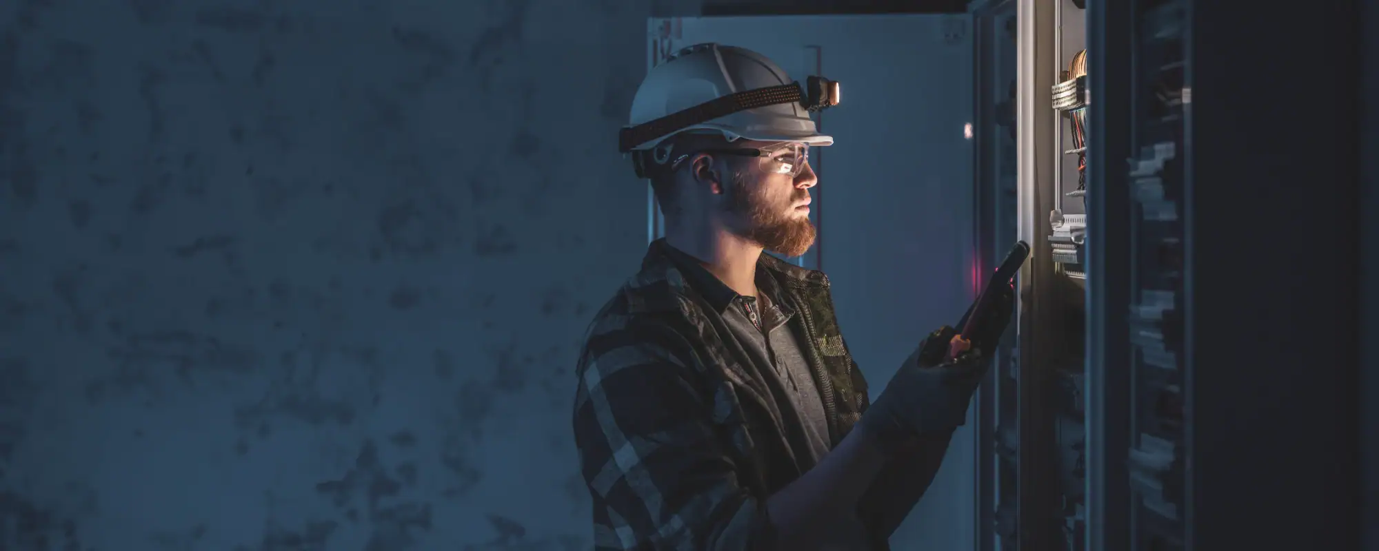 A man wearing a hard hat, safety glasses, and gloves inspects electrical equipment in a dimly lit room. As an electrician in Cook and Will County, he relies on his headlamp and the panel's glow to ensure safe Electrical Services Cook and Will County.