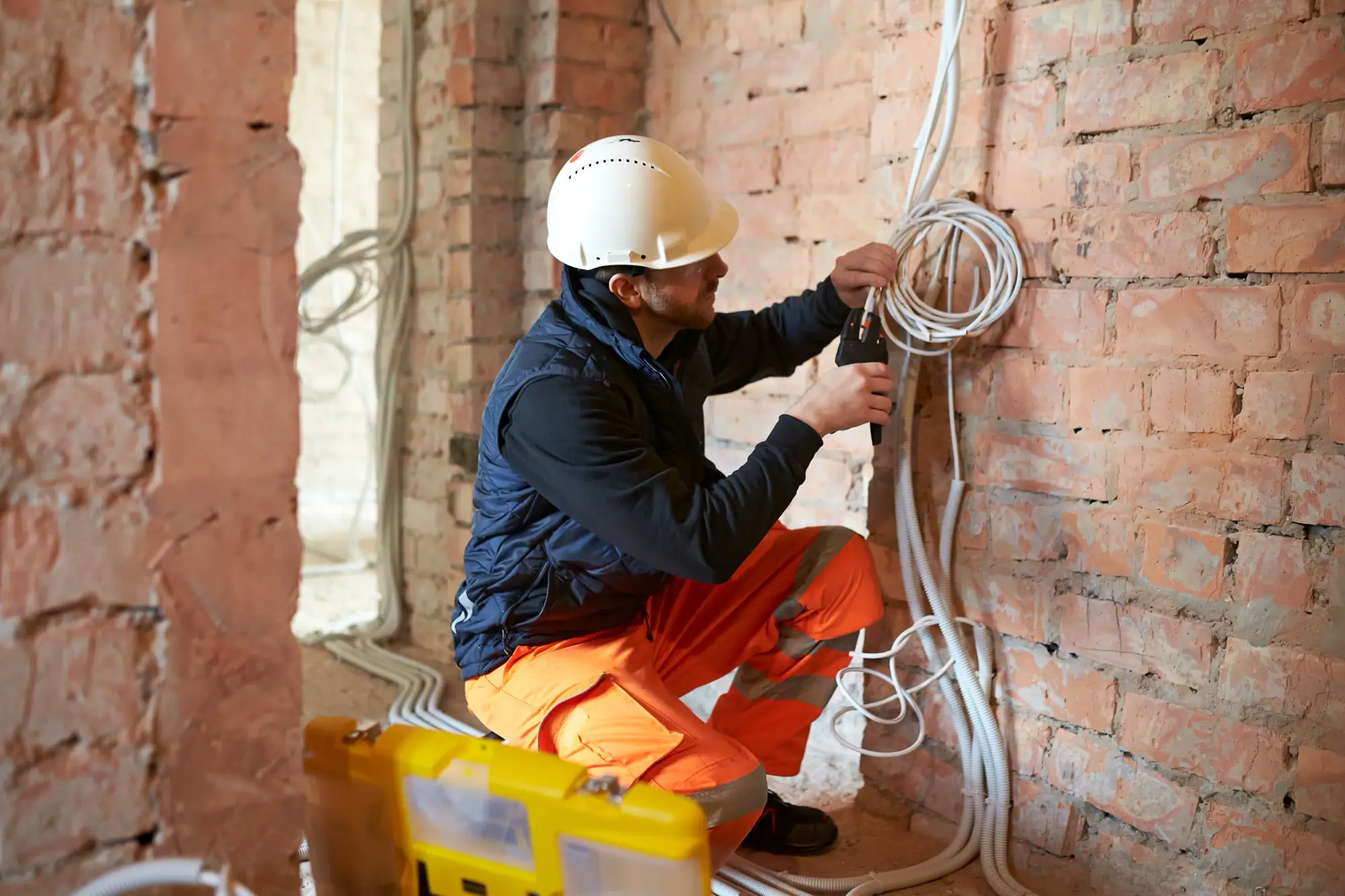 An electrician Cook and Will County, wearing a white hard hat and orange pants, installs or repairs electrical wiring on a bare brick wall inside a building under construction. A yellow toolbox is on the floor nearby.