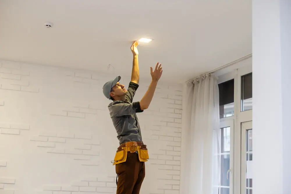 An electrician providing Electrical Services in Cook and Will County, IL, stands on the floor, wearing a cap and tool belt, reaching up to adjust or install a ceiling light fixture in a bright, modern room.