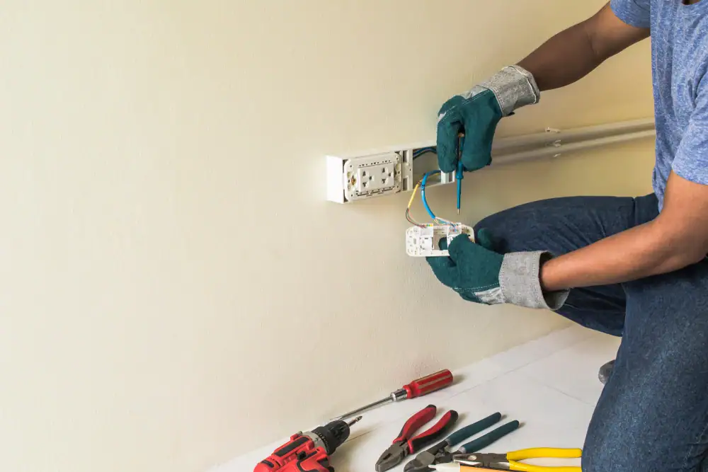 A person wearing gloves installs or repairs a wall electrical outlet using tools laid out on the floor, demonstrating the expertise of an electrician Cook and Will County, IL residents can rely on.