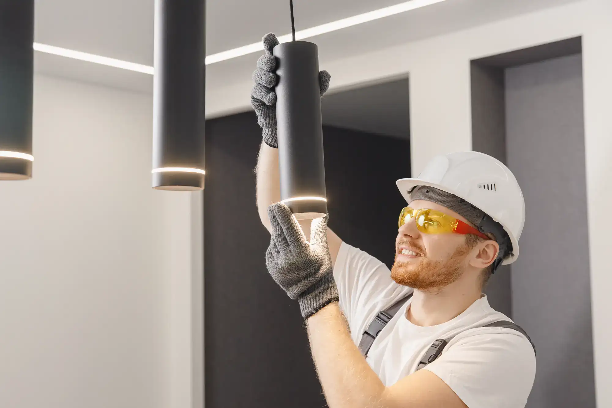 A male electrician in Chicago, IL wearing a white hard hat, yellow safety glasses, gloves, and a white shirt installs or checks a black ceiling light fixture in a bright, contemporary room—showcasing expert Electrical Services
