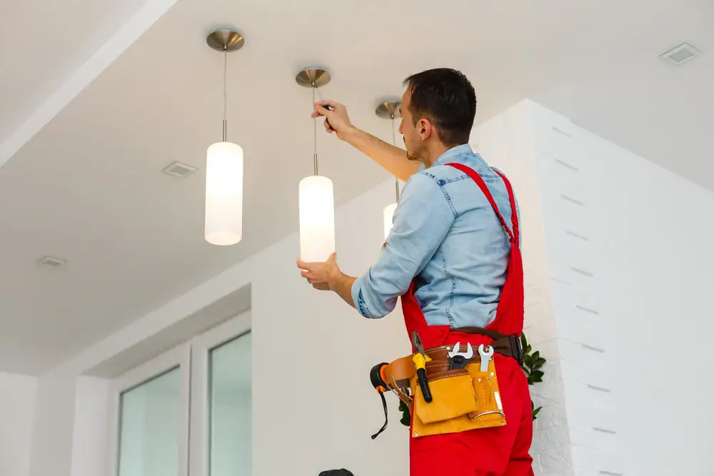 An electrician in red overalls and a tool belt is standing on a ladder, installing or repairing a hanging light fixture on a white ceiling in a modern IL home—quality Electrical Services Cook and Will County.