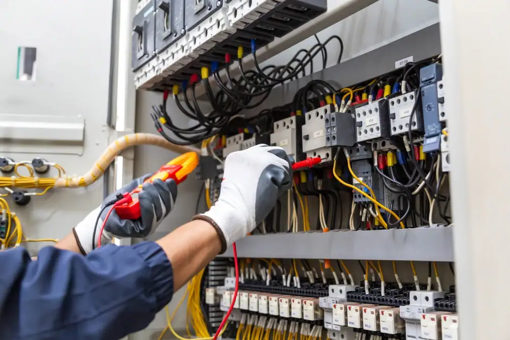 A person wearing gloves uses a multimeter to test electrical wiring inside a control panel filled with cables, switches, and circuit breakers—typical tasks for an electrician Cook and Will County, IL.