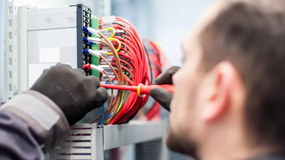A person wearing black gloves uses a screwdriver to work on a panel filled with colorful electrical wires and connections, demonstrating the expertise of an electrician Cook and Will County, IL.