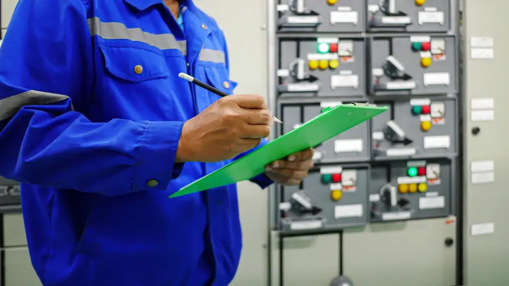 An electrician in Cook and Will County wearing a blue uniform holds a green clipboard and pen, recording information in front of an industrial control panel with switches and indicator lights.