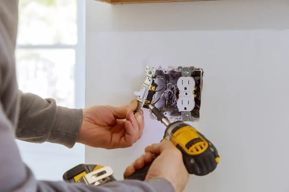 A person, likely an electrician Cook and Will County, IL, uses a yellow power drill to install or repair an electrical outlet and light switch on a white wall, with exposed wiring visible inside the electrical box.