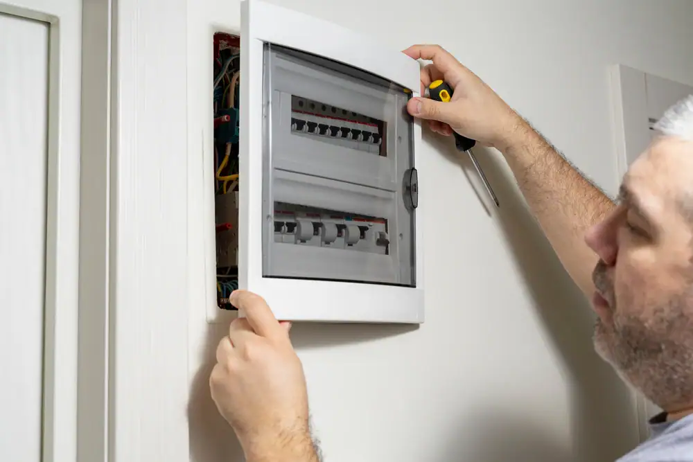 An electrician Cook and Will County, IL opens the door of an electrical breaker panel mounted on a wall, exposing circuit breakers and wiring, while holding a screwdriver.
