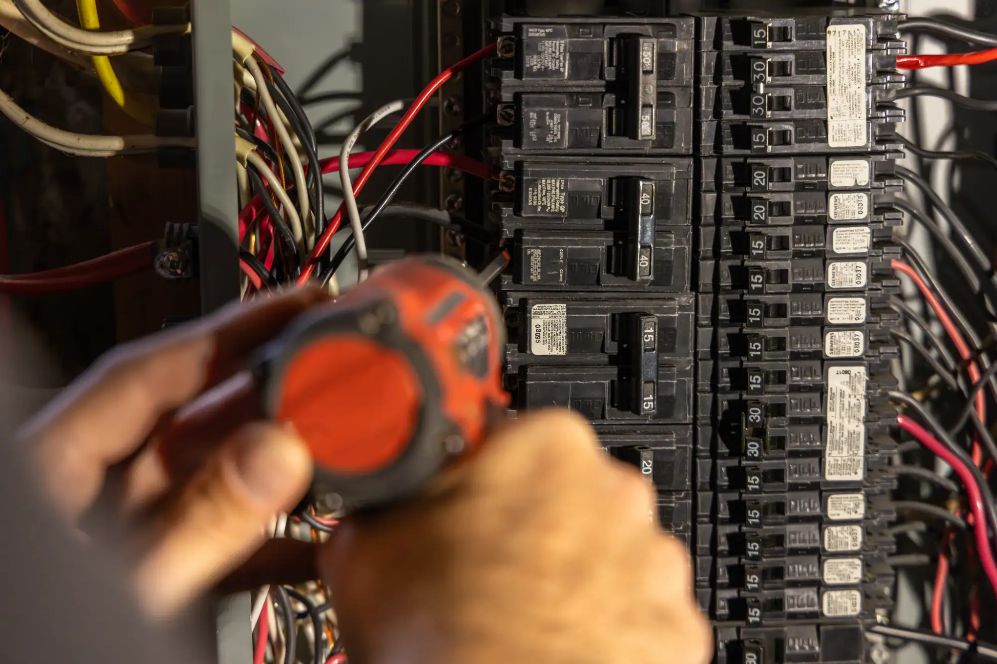 An electrician Cook and Will County, IL uses an orange cordless drill to work on a residential electrical breaker panel filled with wires and circuit breakers.