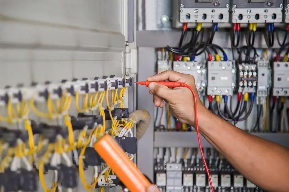 An electrician Cook and Will County uses a multimeter to test electrical connections inside an open control panel filled with yellow wires, switches, and circuit components.