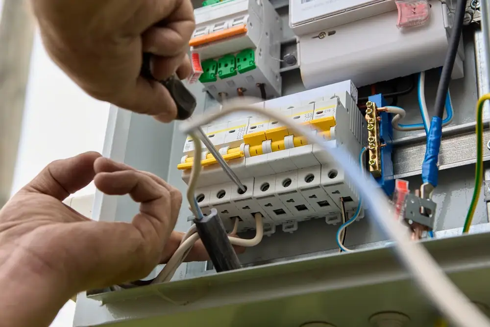 Close-up of hands installing or repairing electrical wiring inside a circuit breaker panel, using a screwdriver—perfect for showcasing the expertise of an electrician Cook and Will County, IL. Different colored wires connect to switches and terminals.