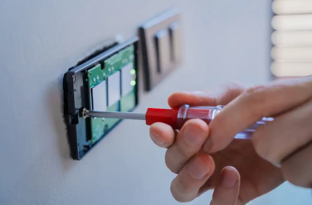 Close-up of an electrician Cook and Will County, IL, using a screwdriver to install or repair a light switch on a wall, with exposed wiring and circuit components visible.