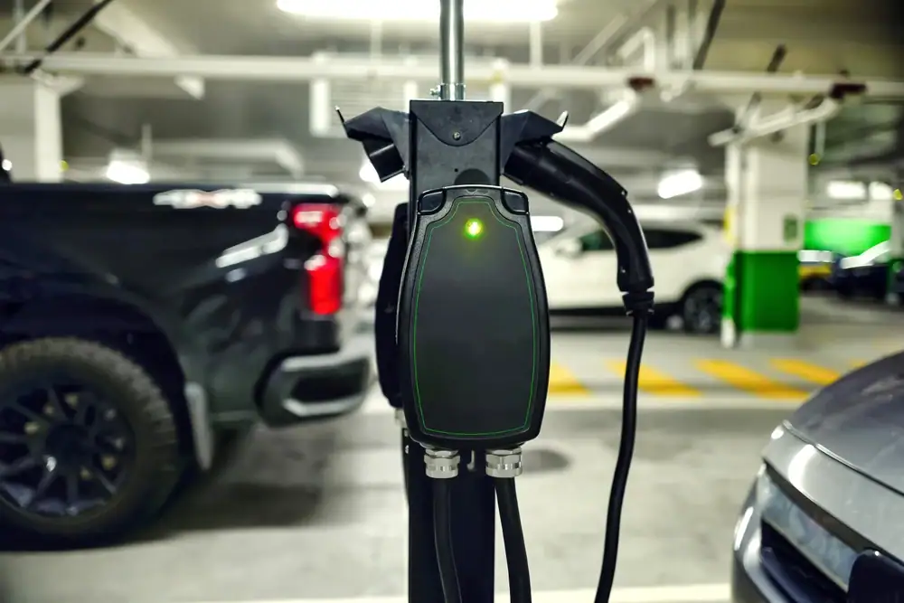 A close-up of an electric vehicle charging station with a glowing green light, located in an IL parking garage where cars are parked nearby—perfect for those seeking Electrical Services Cook and Will County.