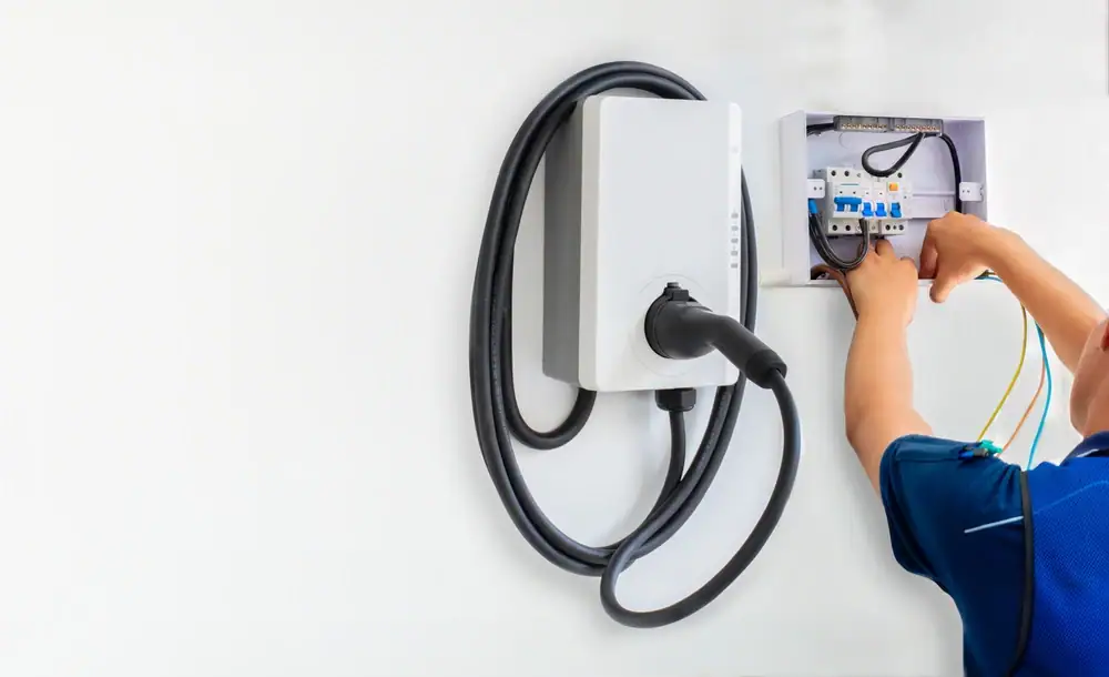 An electrician Cook and Will County installs or repairs an electric vehicle charging station, connecting wires inside an open electrical panel mounted on a white wall. A charging cable is coiled on the station.