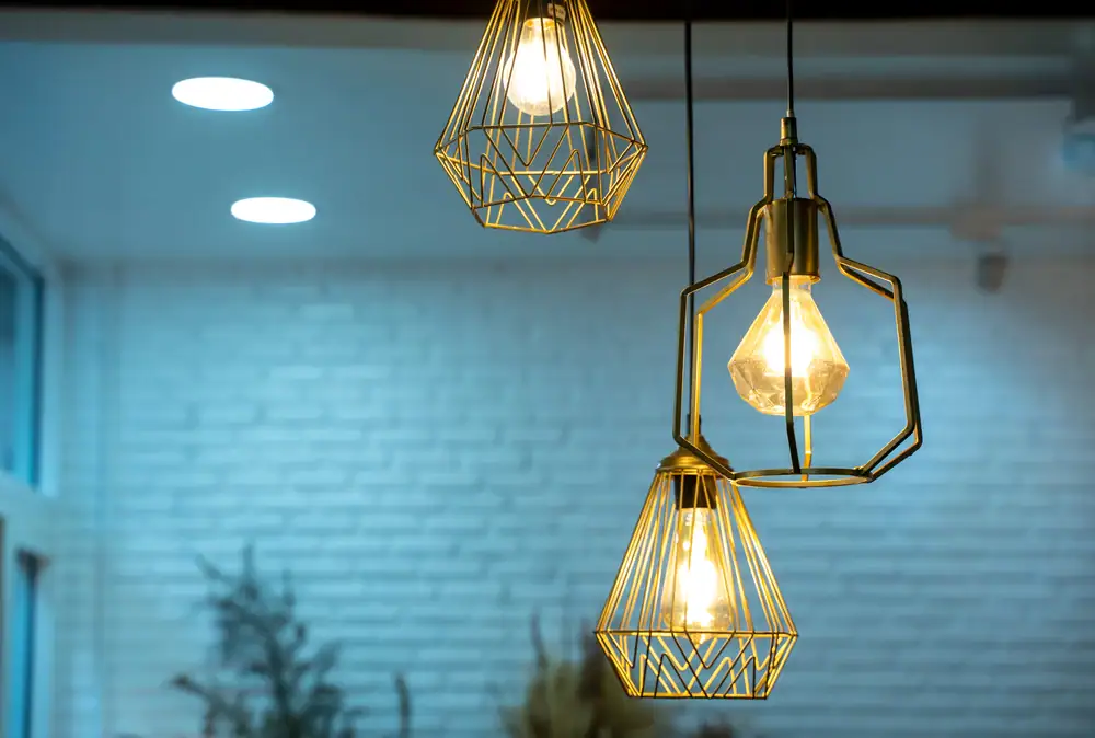 Three geometric pendant lights with exposed bulbs hang from the ceiling in front of a white brick wall, beautifully illuminated—perfect lighting designed by expert Electrical Services Cook and Will County, IL.
