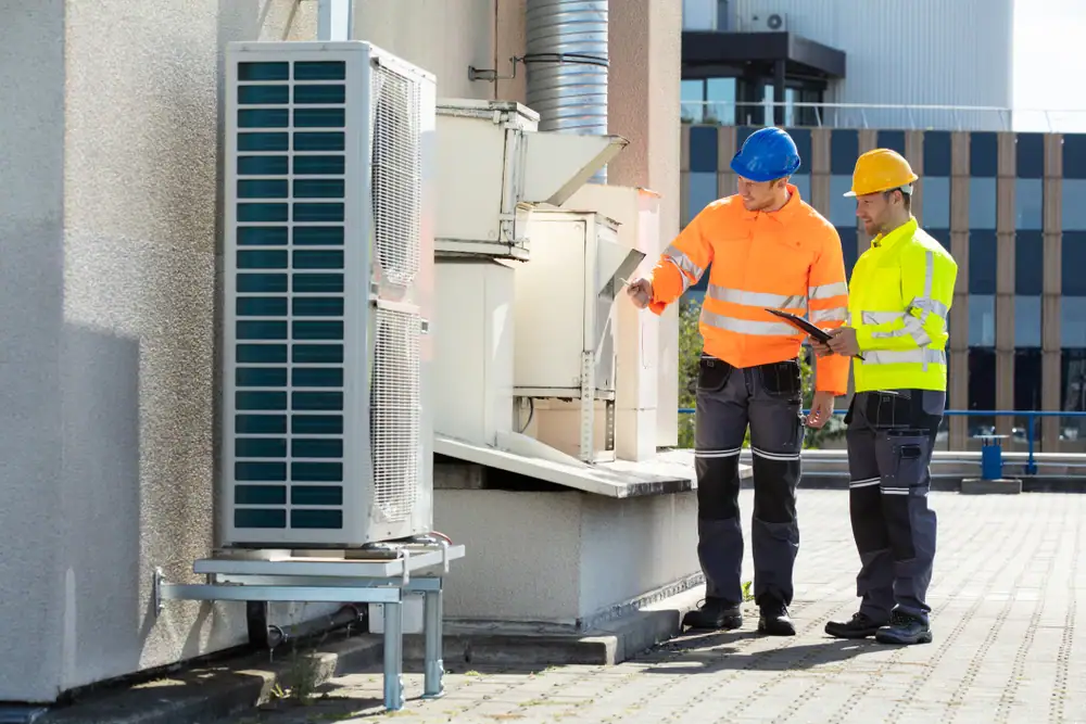 Two workers in safety gear and helmets stand on a rooftop, inspecting and discussing an HVAC unit. An electrician Cook and Will County holds a clipboard while the other points to the unit, ensuring all electrical services are up to code.