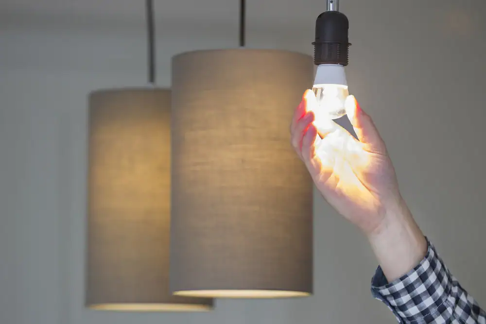 A person with a plaid sleeve is installing a lit LED bulb in a ceiling lamp with a fabric lampshade, while another similar fixture is visible behind—ideal for those seeking Electrical Services Cook and Will County, IL.