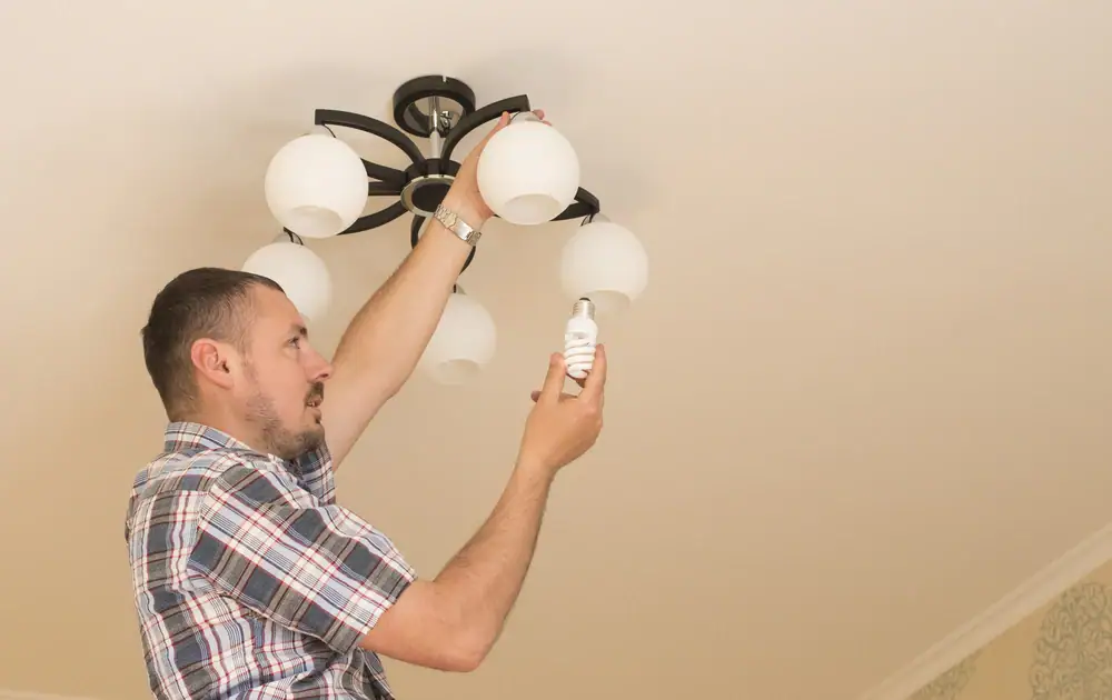 A man in a plaid shirt is standing on the floor, changing a light bulb in a ceiling chandelier. For safe and reliable help, contact an electrician Cook and Will County for expert electrical services.