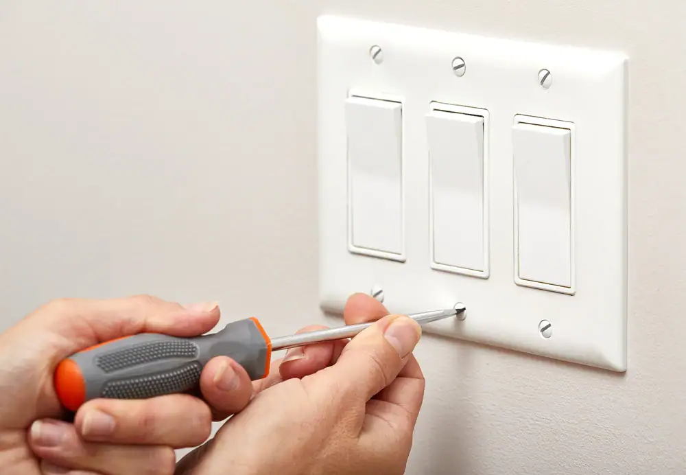 An electrician Cook and Will County, IL uses a screwdriver to tighten a screw on a white wall plate covering three light switches mounted on a light-colored wall.