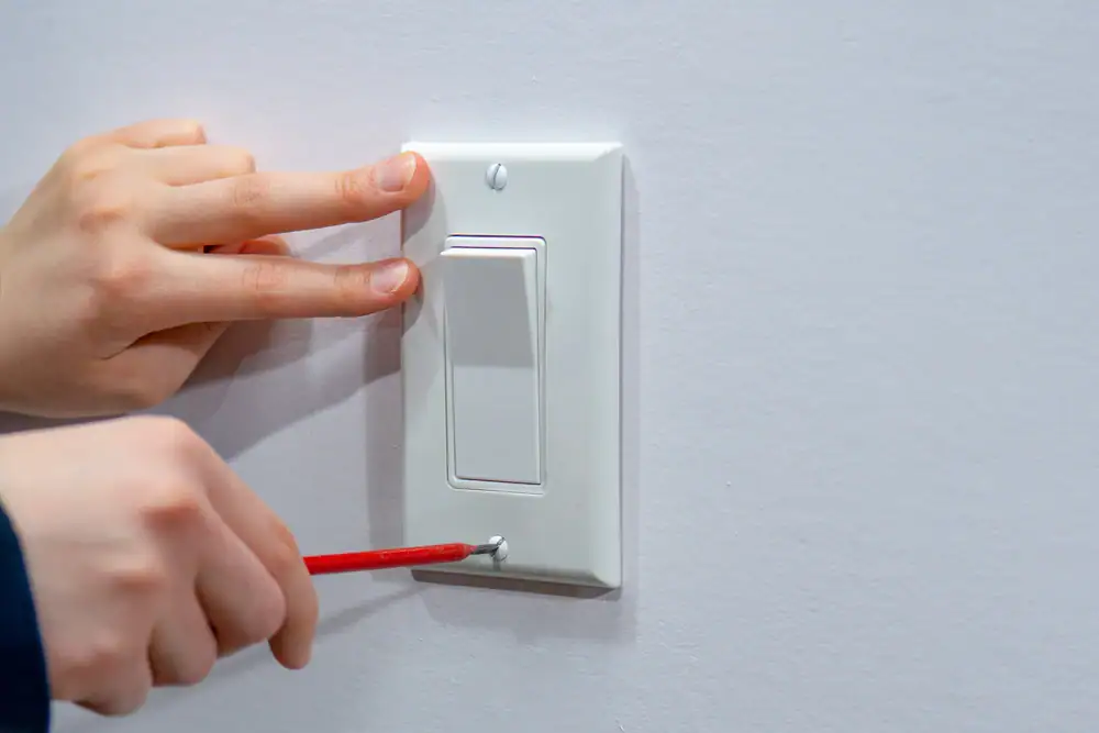 A person, possibly an electrician Cook and Will County, uses a screwdriver to loosen the screw on the bottom of a white light switch plate, holding it steady with one hand—showing typical Electrical Services Cook and Will County, IL.