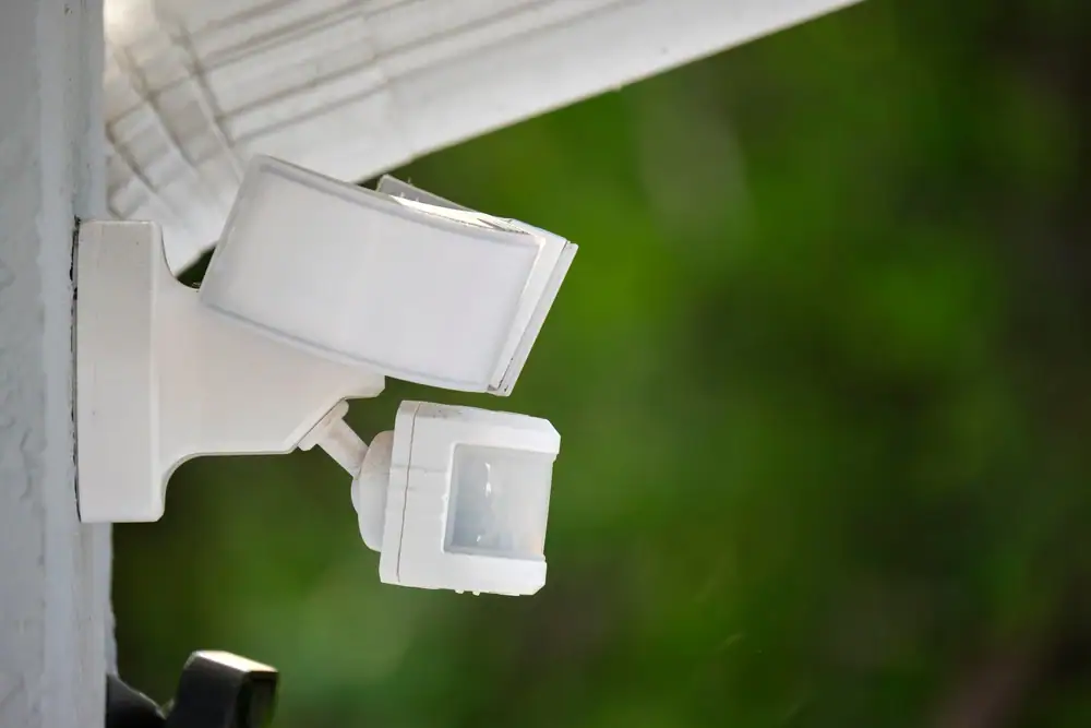 A white motion sensor security light is mounted on an exterior wall, with a blurred green background suggesting outdoor foliage—ideal for homes in IL who trust Electrical Services Cook and Will County for safety solutions.