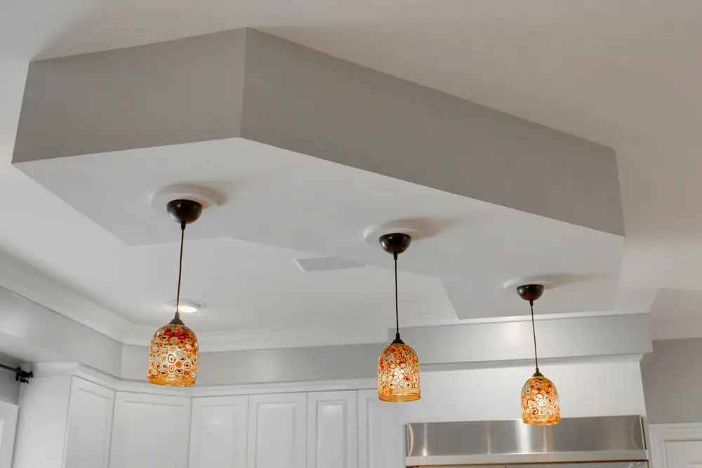Three pendant lights with patterned orange glass shades hang from the ceiling, each suspended over a kitchen area with white cabinetry and a unique geometric ceiling design by an expert electrician Cook and Will County, IL.