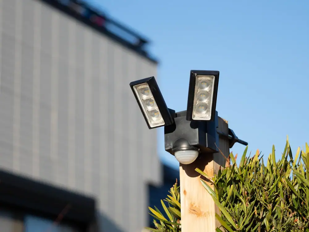 A security floodlight with two rectangular LED lamps, expertly installed by an electrician in Cook and Will County, mounted on a wooden post near green bushes, set against a clear blue sky and a modern building background.