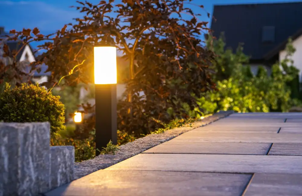 A glowing outdoor garden lamp illuminates a stone pathway at dusk, showcasing the quality of Electrical Services in Cook and Will County, IL, surrounded by greenery and residential houses in the background.