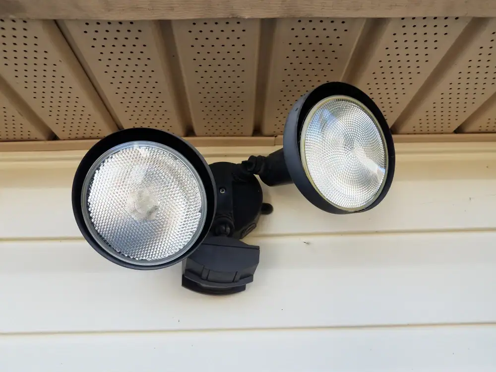 A close-up of an outdoor motion sensor floodlight with two round lamps, expertly installed by an electrician Cook and Will County, mounted on the exterior wall under the eaves. The background shows beige siding and perforated soffit panels.