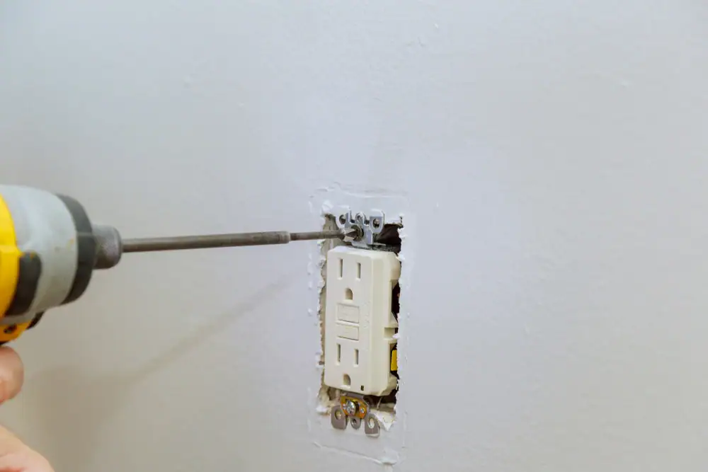 An electrician Cook and Will County uses a power drill to install or remove a screw from a white electrical outlet set in a wall with uneven paint or plaster around the socket.